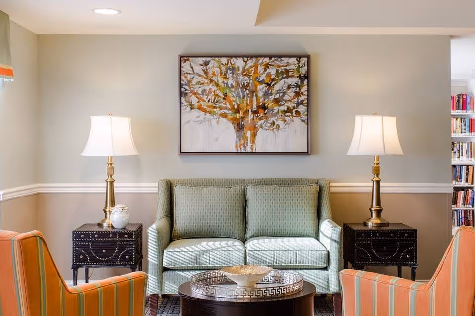 A cozy living room area with a light green patterned sofa centered between two dark wooden side tables, each topped with a brass lamp with white shades. Two orange striped armchairs face the sofa. Above the sofa hangs a large framed painting of a tree with autumn-colored leaves. A round coffee table with a decorative bowl is in front of the sofa. A bookshelf filled with books is visible on the right side of the image.