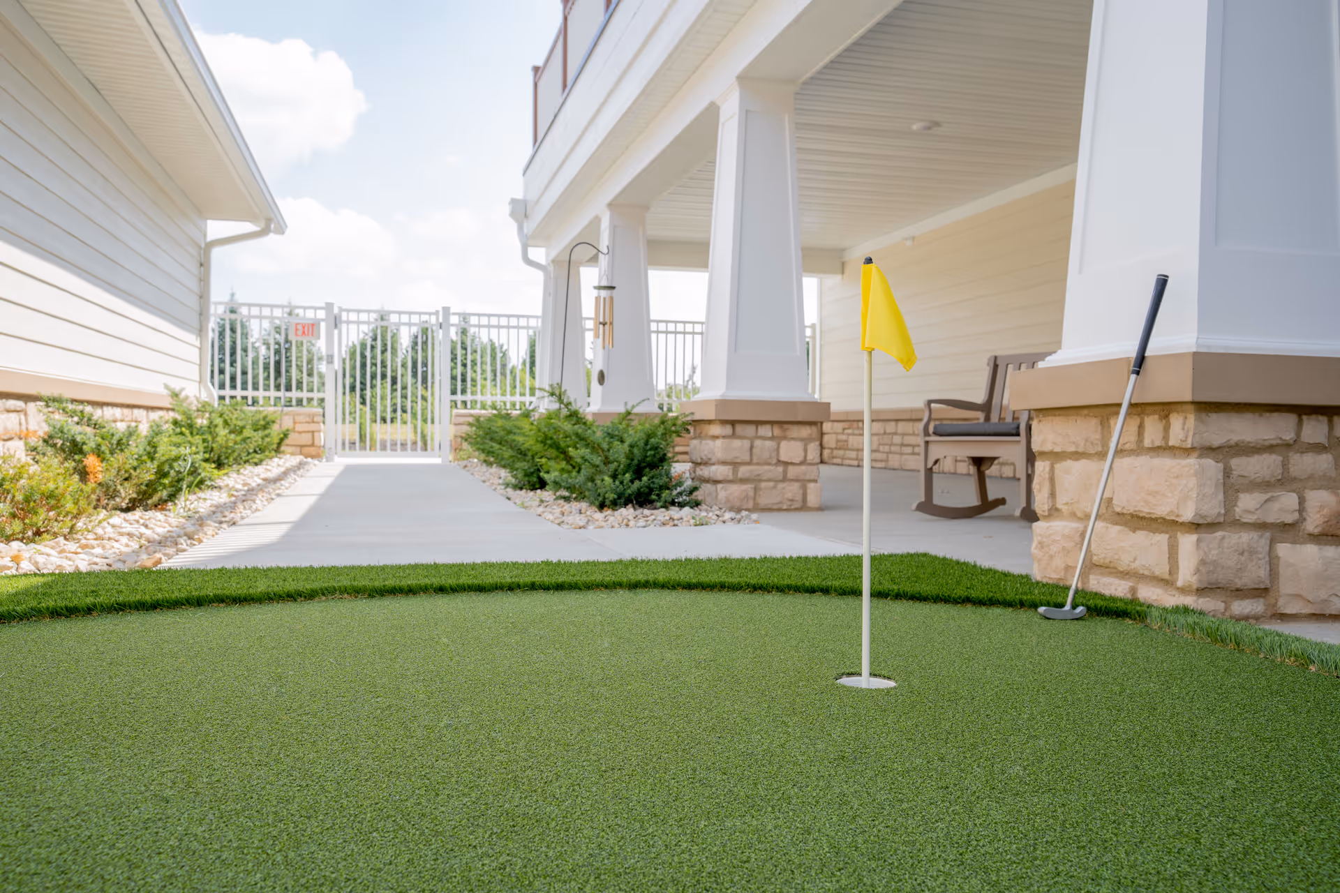 A small outdoor putting green with a yellow flag and a golf putter leaning against a stone pillar. The area is adjacent to a covered patio with a bench and surrounded by landscaping with bushes and rocks. A white fence and gate are visible in the background under a partly cloudy sky.