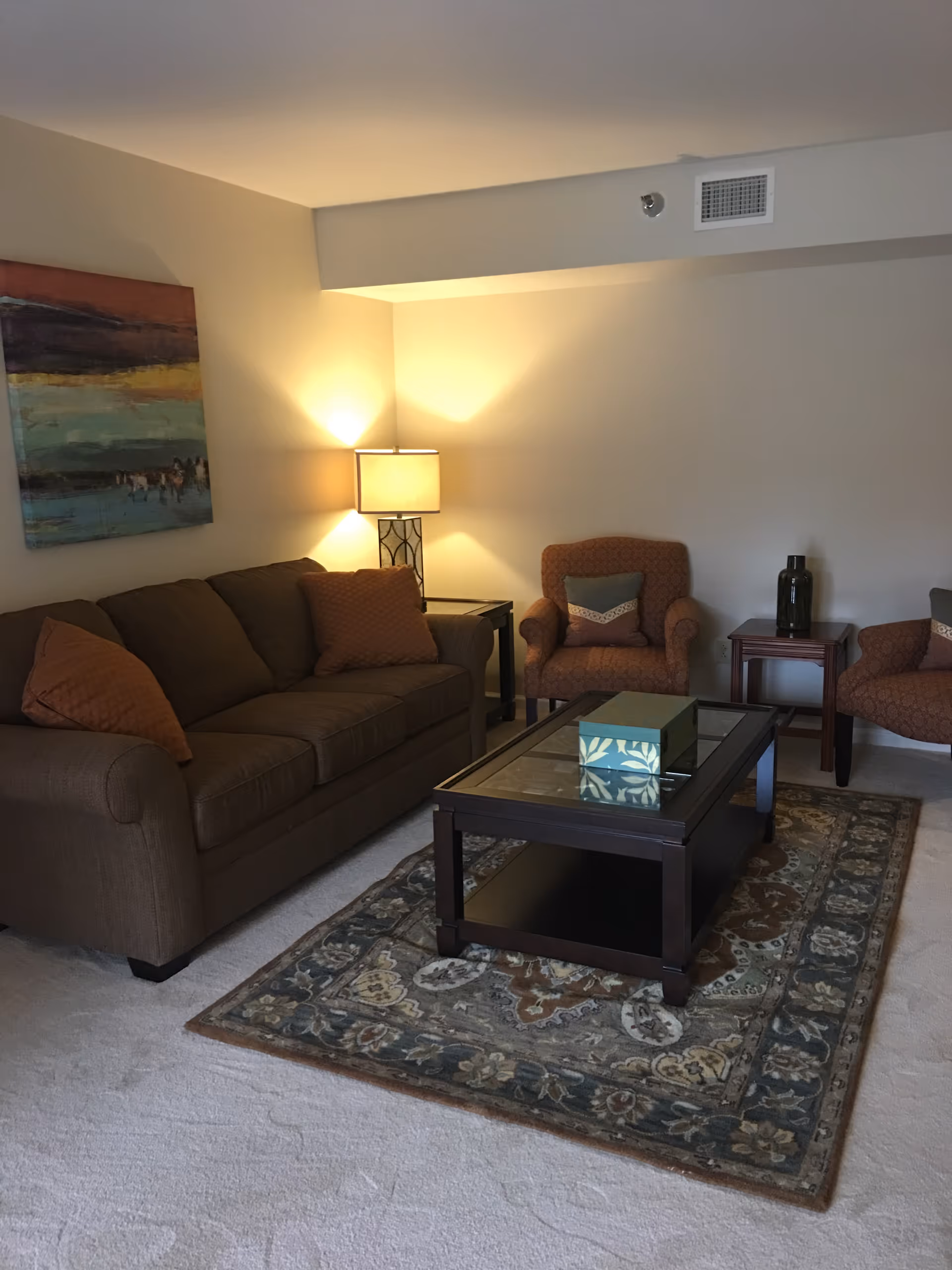 A cozy living room with a brown sofa and two matching armchairs with patterned cushions. A wooden coffee table with a glass top sits on a decorative area rug. A table lamp on a side table illuminates the corner, and a colorful abstract painting hangs on the wall.