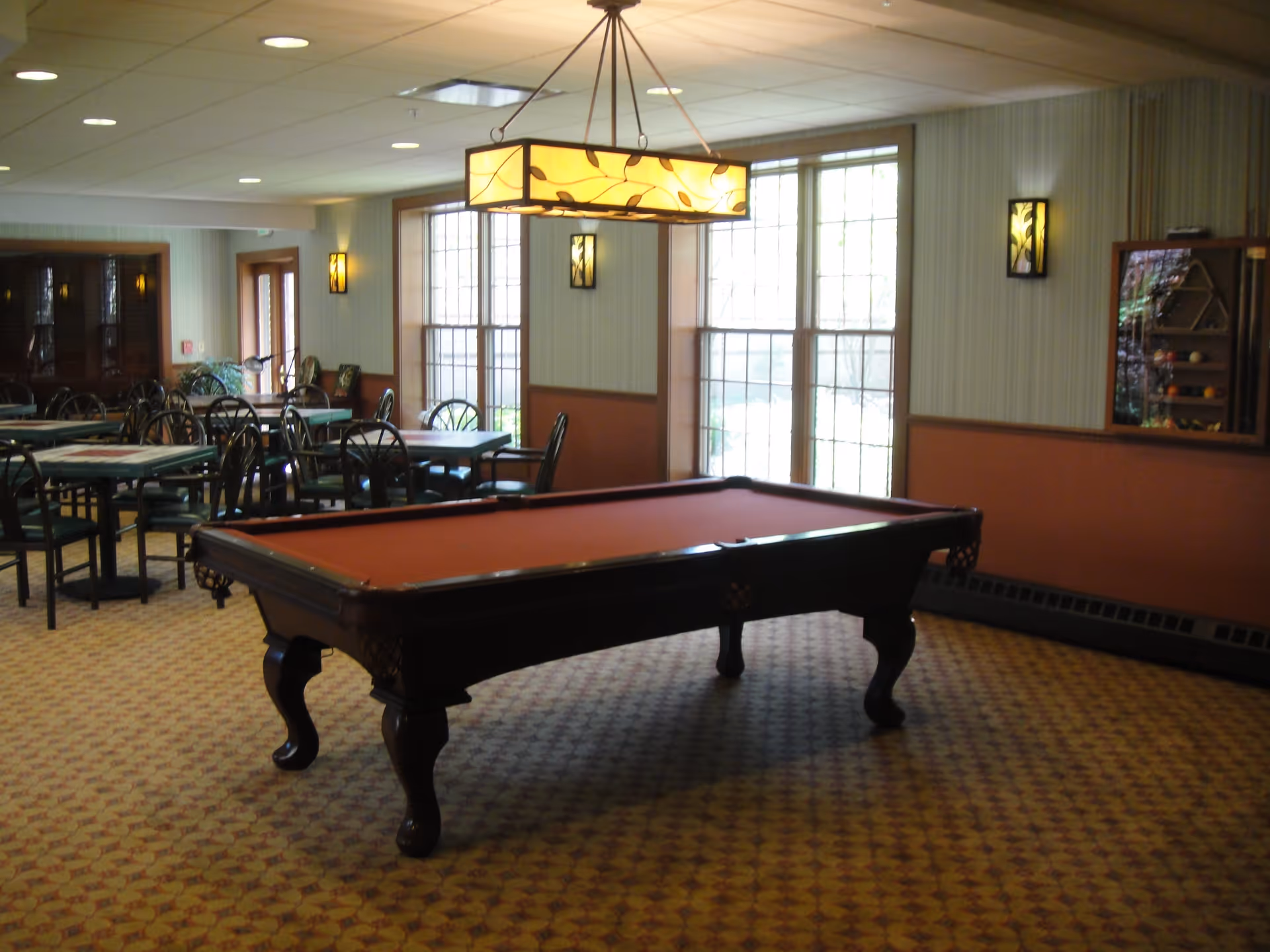 Interior room with a pool table in the foreground and several tables with chairs in the background. The room has large windows letting in natural light, patterned carpet, and decorative wall sconces. A rectangular hanging light fixture is above the pool table.