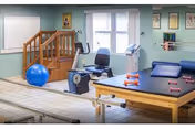 Bright therapy/rehab room with parallel walking bars, a recumbent bike, therapy table, exercise ball, small dumbbells and a practice staircase under windows.