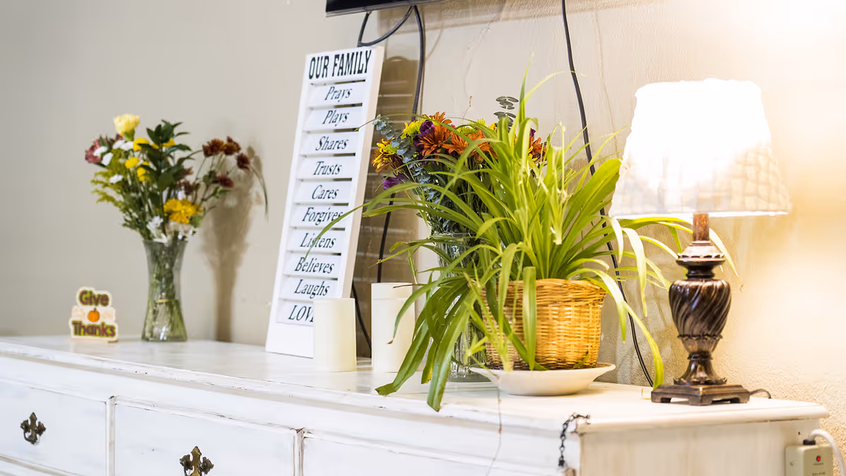 White sideboard topped with a lamp, potted plants, flower vases and a decorative 'Our Family' sign.