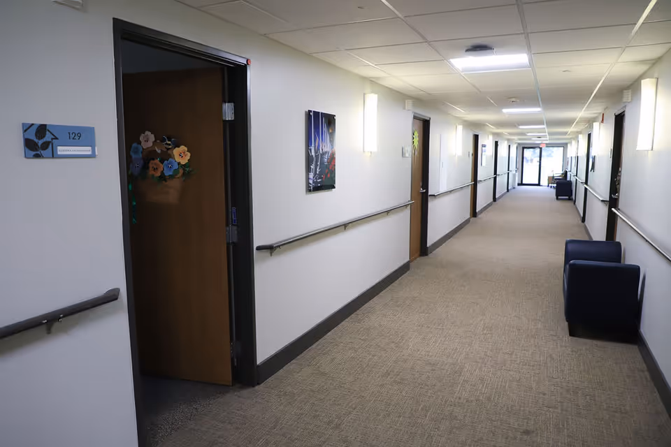 A long, well-lit hallway in an assisted living facility with beige carpet and white walls. Several closed wooden doors line the hallway, one door is partially open and decorated with colorful paper flowers. Handrails run along both sides of the hallway, and there are a few dark blue armchairs placed against the walls. At the far end, glass doors lead to another area with natural light coming through.