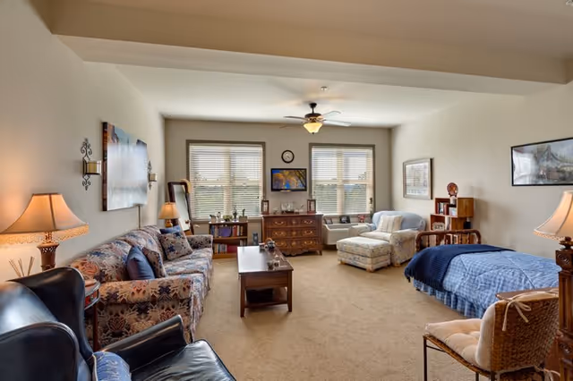 A spacious senior living room with a floral patterned sofa, a blue armchair, a wooden coffee table, and a bed with a blue blanket. The room has beige carpet, two large windows with blinds, a ceiling fan with light, and various lamps and framed pictures on the walls. There is also a wooden dresser, a cushioned armchair with an ottoman, and a wicker chair.