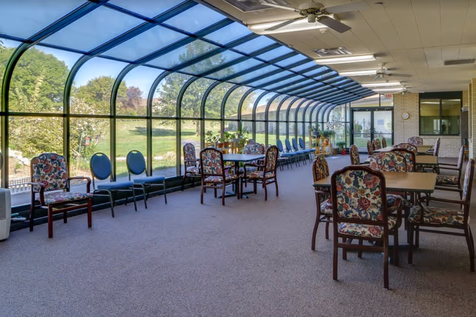 Sunlit solarium-style common dining area with arched glass windows, multiple tables and floral-upholstered chairs overlooking a lawn.