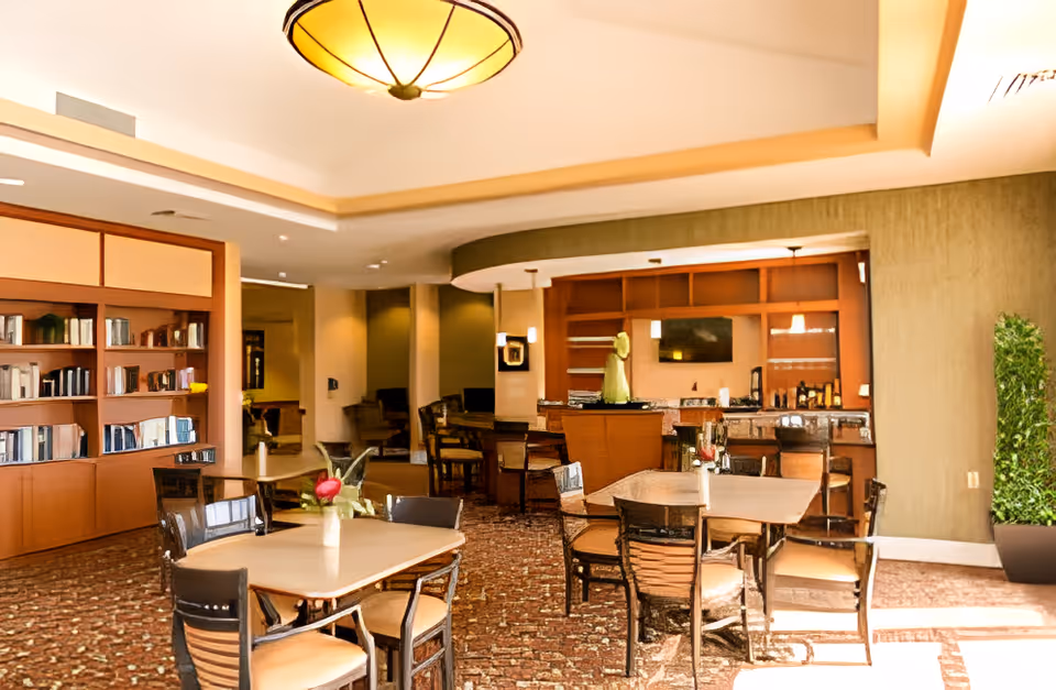 Bright communal dining and lounge area with tables, chairs, bookshelves and a serving counter.