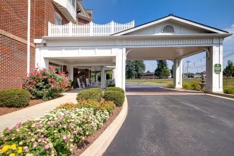 Entrance area of Harbour Assisted Living facility showing a covered driveway with white pillars and a white picket fence on top. There are colorful flower beds and green bushes along the sidewalk next to a red brick building. The sky is clear and blue.