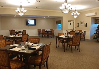 A spacious dining room with multiple wooden tables and cushioned chairs arranged neatly. Each table is set with white cups, plates, and napkins. The room is well-lit with chandeliers hanging from the ceiling. A television is mounted on the far wall, and there are framed pictures on the walls. A decorated Christmas tree is visible in the corner, indicating a festive season.