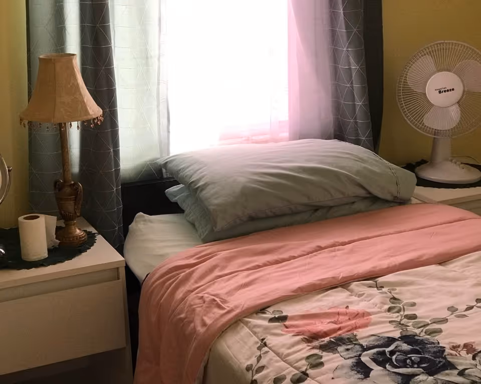 A neatly made bed with pink and floral bedding, stacked pillows, a bedside lamp and a fan beside a curtained window.