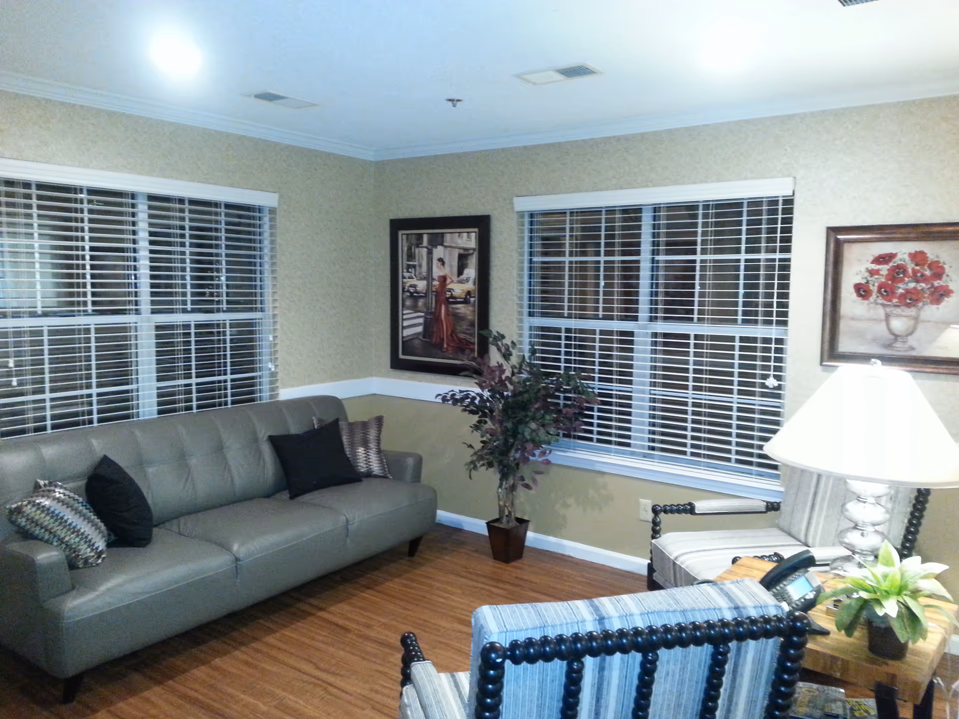Comfortable living room with a gray sofa, striped armchairs, potted plant, framed artwork, and a lamp in front of two large windows with blinds.