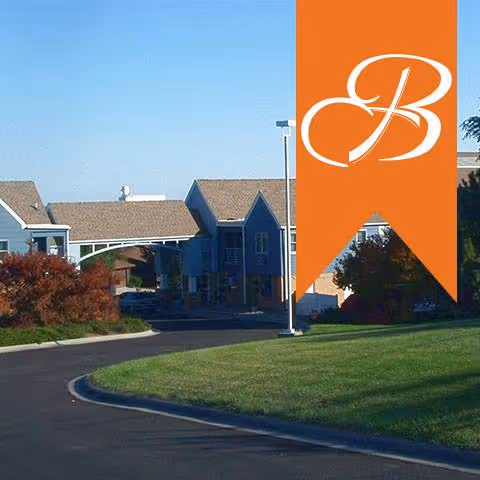 Front exterior of a two-story assisted living building with a curved driveway, lawn, and an orange banner showing a white stylized 'B'.