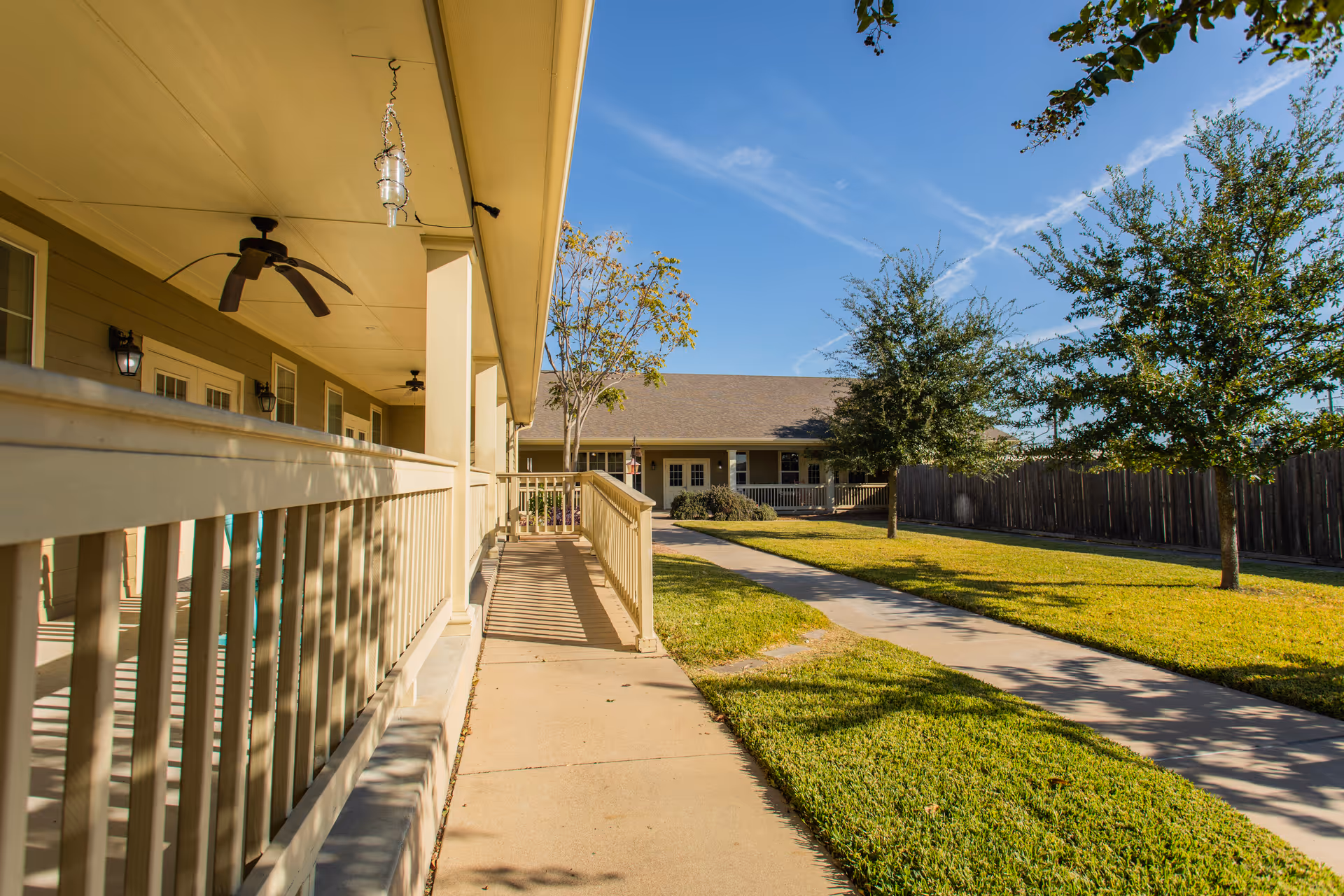 Outdoor view of a senior living facility showing a paved walkway alongside a porch with ceiling fans and hanging lanterns. The porch is bordered by a beige railing, and the walkway is flanked by well-maintained grass and trees under a clear blue sky.