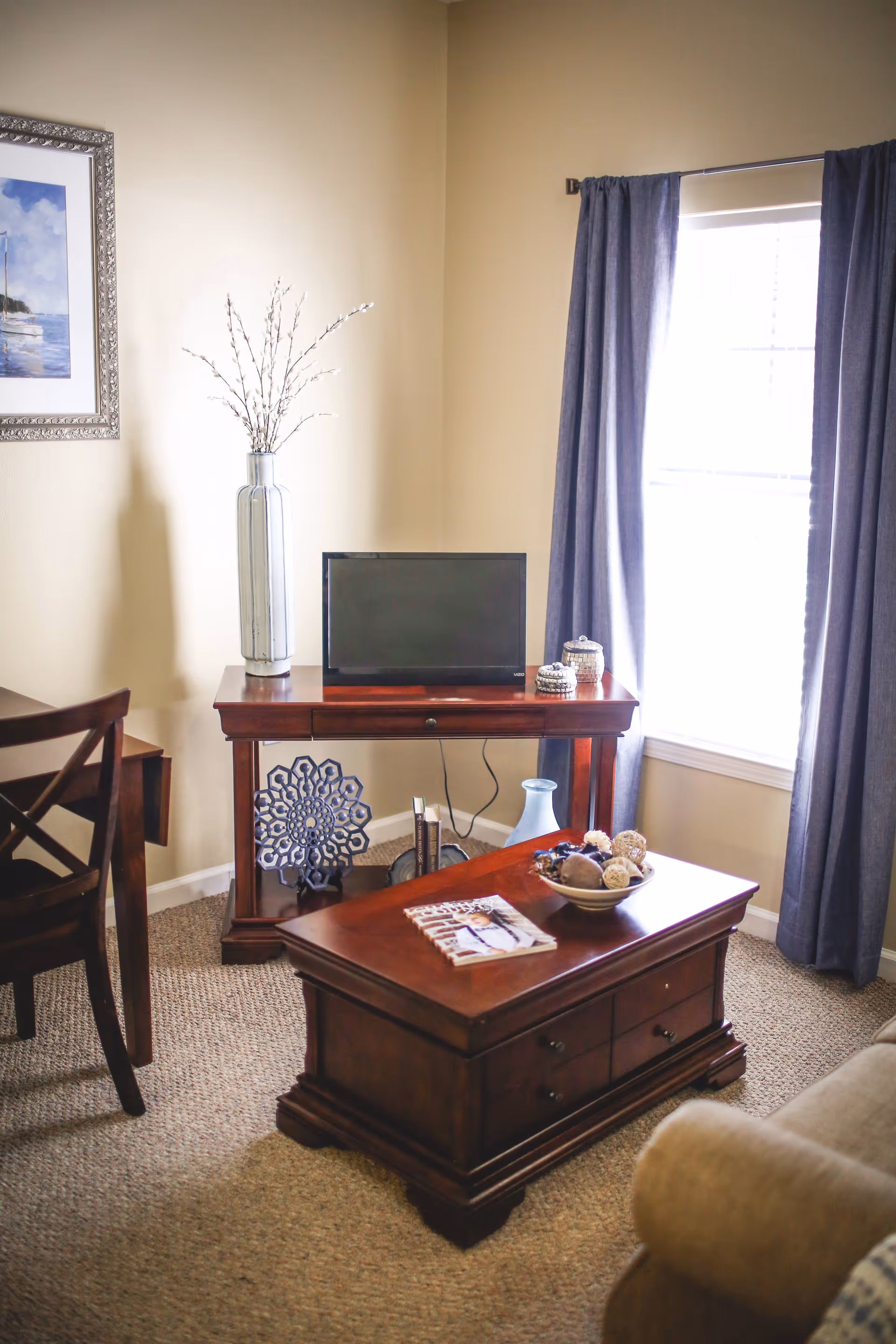 A cozy living room corner with a wooden coffee table holding a decorative bowl and a magazine, a wooden TV stand with a flat-screen TV, decorative items including a tall vase with branches, and a window with blue curtains letting in natural light.