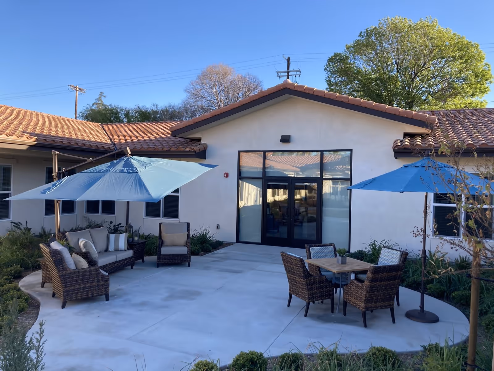 Outdoor patio area at The Preserve at Woodland Hills Assisted Living & Memory Care with wicker seating including a sofa and chairs, two blue umbrellas providing shade, a small table with four chairs, and a building with a tiled roof and large glass doors in the background.
