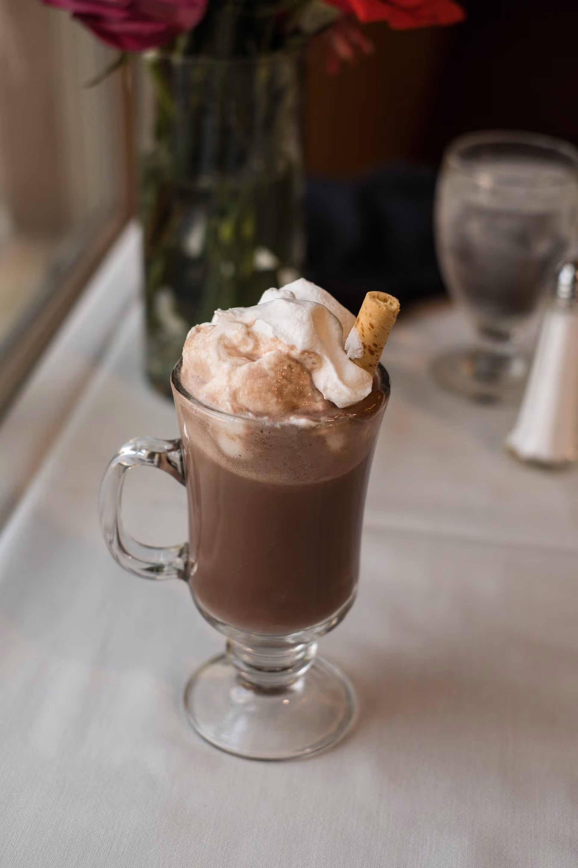 A glass mug filled with hot chocolate topped with whipped cream and a wafer stick, placed on a white tablecloth with a blurred background including a glass of water, salt shaker, and a vase with flowers.