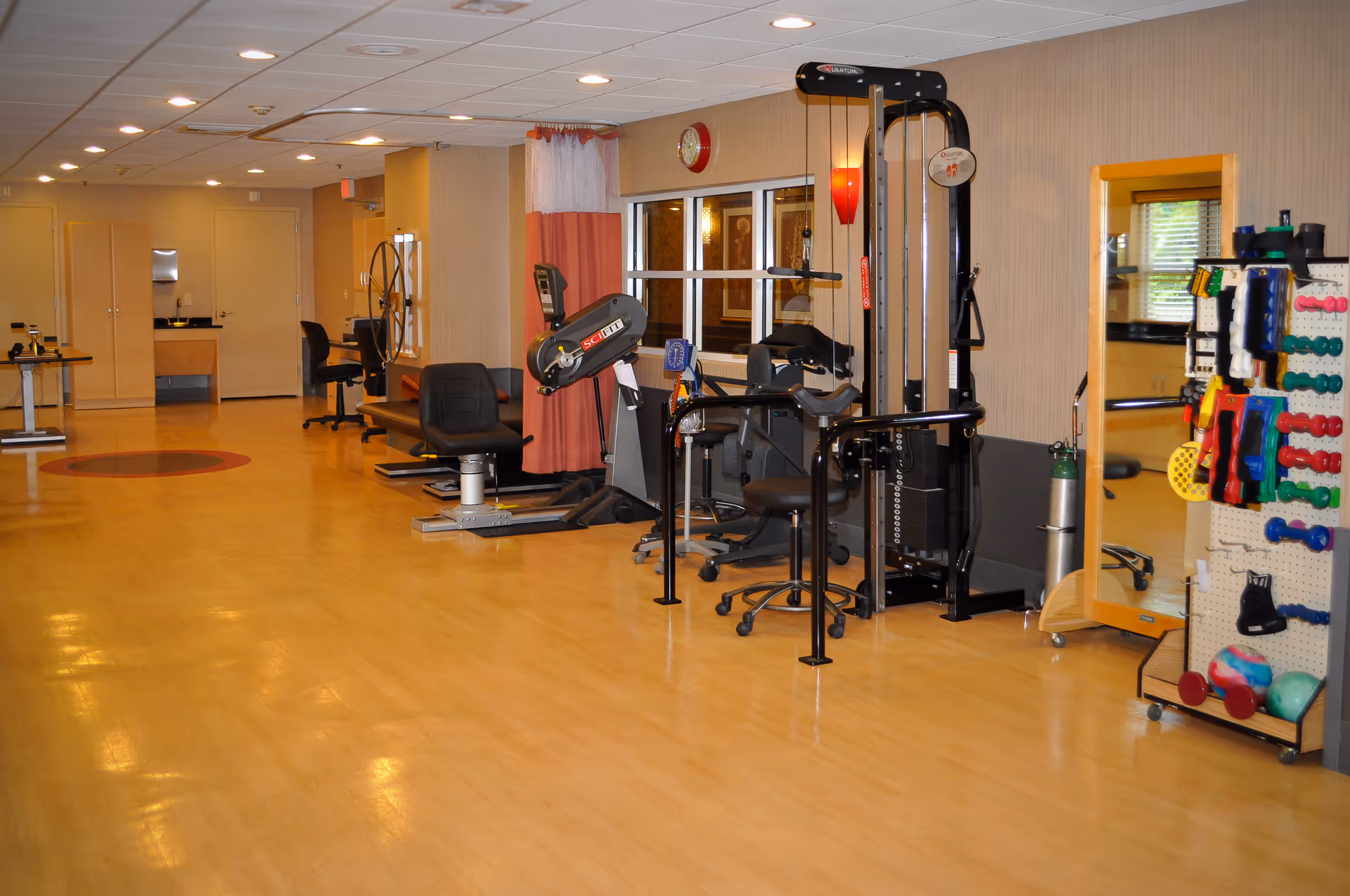 A spacious exercise room in a senior living facility with various fitness equipment including stationary bikes, a multi-function weight machine, and a rack of colorful hand weights and exercise balls. The room has wooden flooring, beige walls, a large mirror, and ceiling lights.