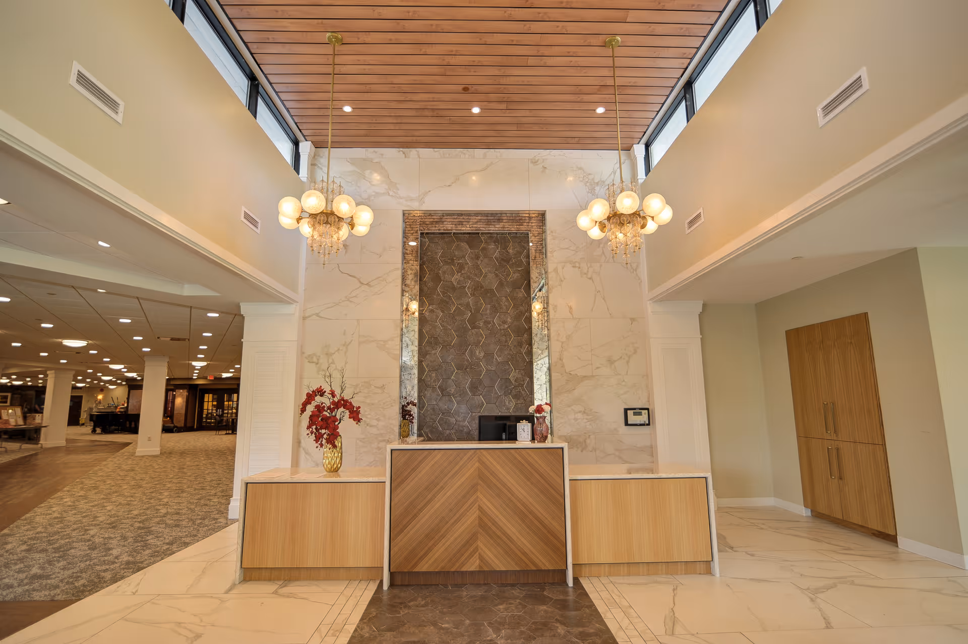 Reception area with a wooden front desk, marble walls, and two hanging chandeliers. There are decorative vases with red flowers on the desk. The ceiling is wooden with recessed lighting, and there is a carpeted hallway leading to other rooms on the left.
