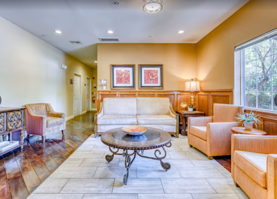Cozy memory care living room with a sofa, armchairs, round coffee table, framed artwork, and a large window.