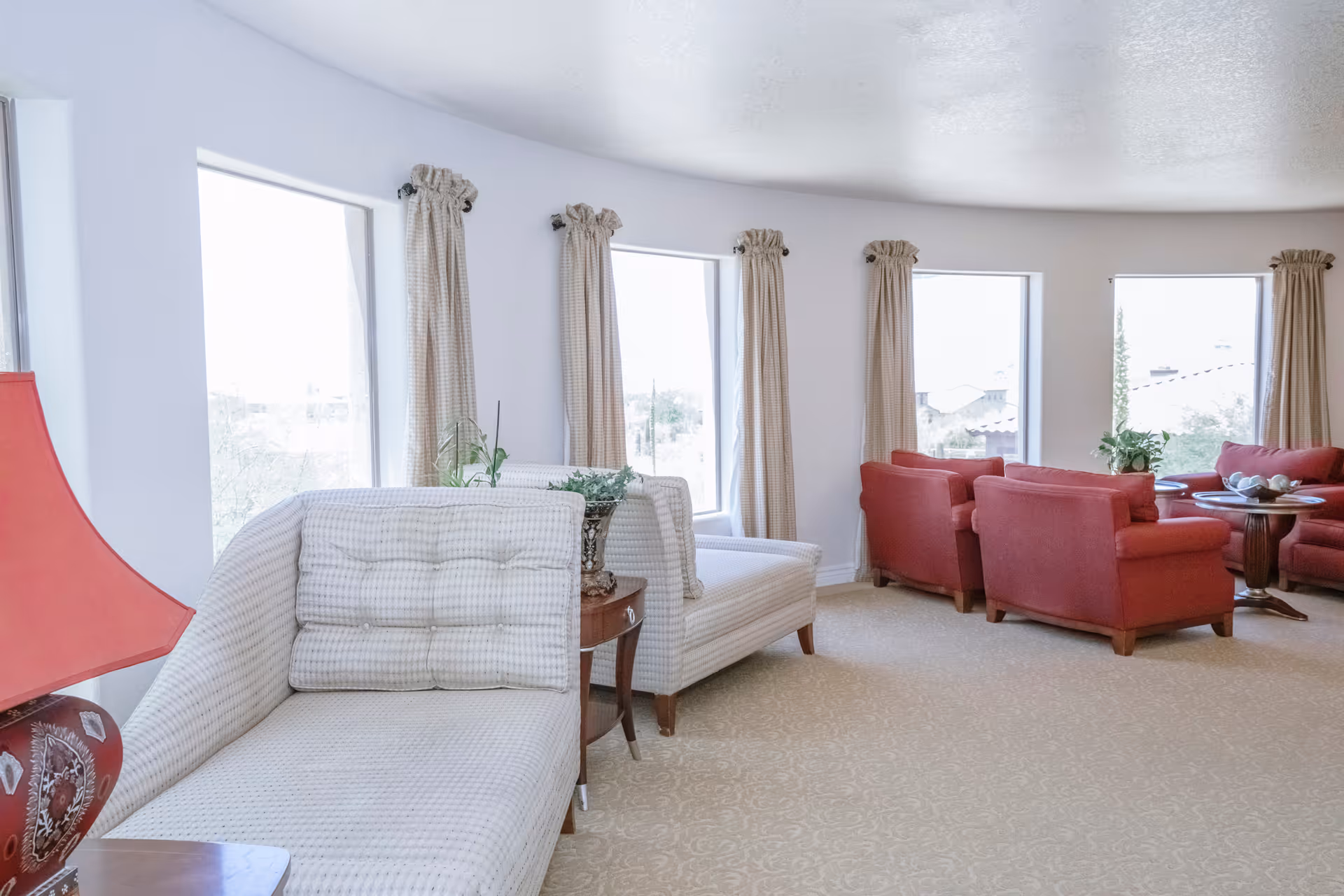 A bright and spacious living room area with large windows covered by beige curtains. The room features a light-colored patterned sofa with matching cushions, a red table lamp on a side table, and several red armchairs arranged around a wooden coffee table with decorative items. The carpet is beige with a subtle pattern.