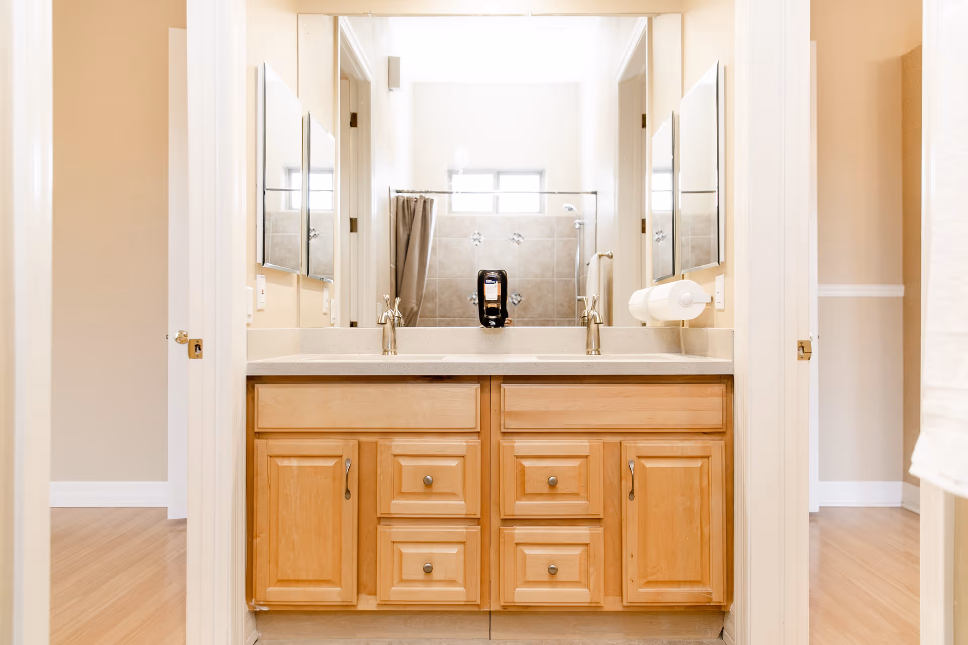 A bathroom vanity with a double sink and wooden cabinets below. Above the sinks are two mirrors, and in the reflection, a shower with a curtain and tiled walls is visible. The bathroom has light-colored walls and a small window above the shower.
