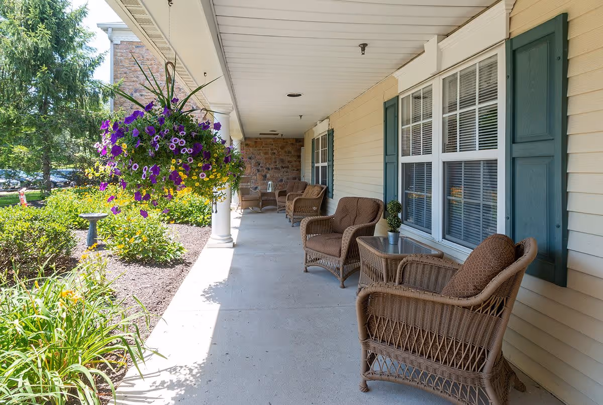 A covered outdoor porch area with wicker chairs and small tables arranged along the wall of a building. There are windows with green shutters on the building side and a hanging basket with purple and yellow flowers. The porch overlooks a garden with green shrubs, flowers, and a birdbath.