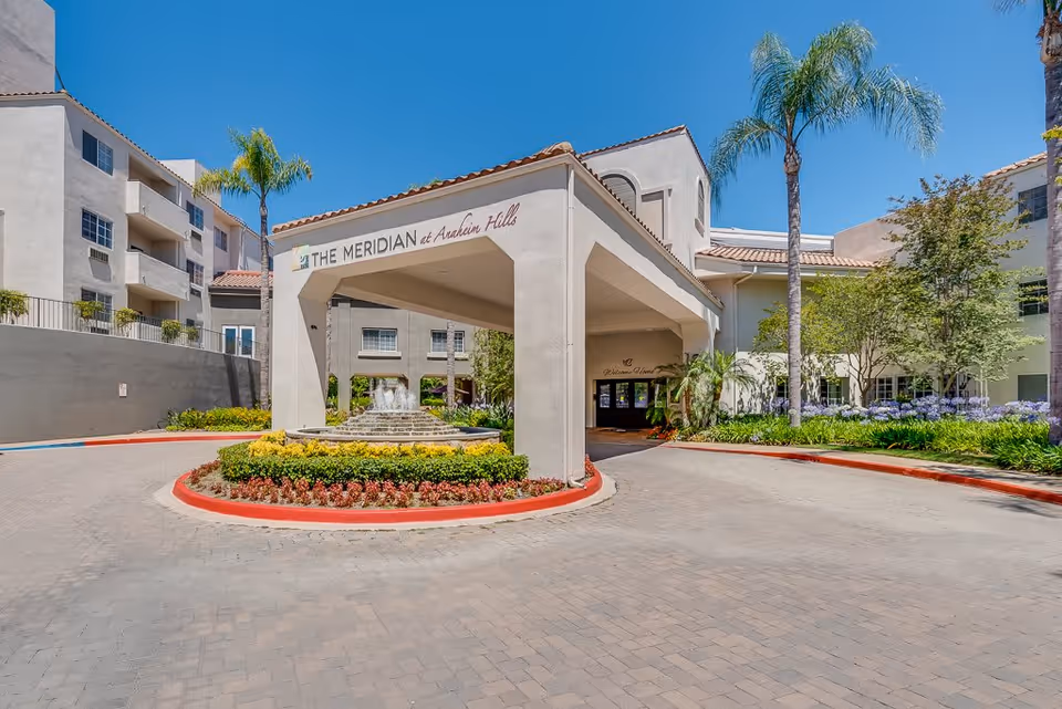 Exterior view of The Meridian at Anaheim Hills senior living facility entrance with a covered drop-off area, landscaped flower beds, palm trees, and a clear blue sky.