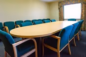 A conference or meeting room with a large oval wooden table surrounded by teal upholstered chairs. The room has beige walls, a window with patterned curtains, and carpeted flooring.