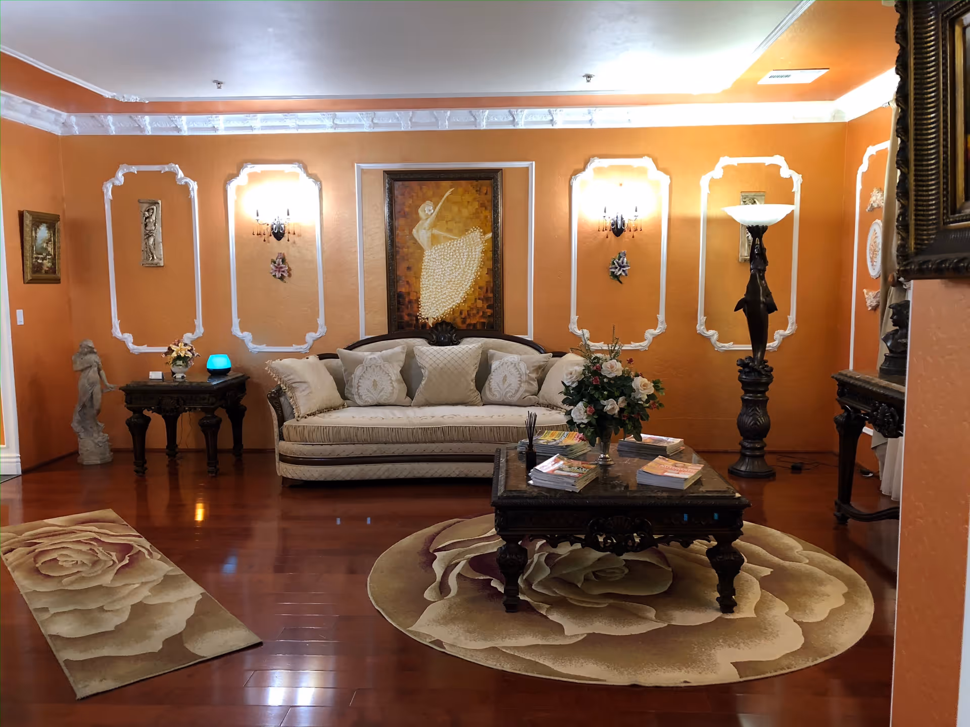 A cozy living room with orange walls and white decorative molding. The room features a cream-colored sofa with multiple cushions, a dark wooden coffee table with magazines and a flower arrangement, a tall floor lamp with a dolphin design, and two rugs with large rose patterns on a polished wooden floor. Various framed artworks and sculptures decorate the walls and corners.
