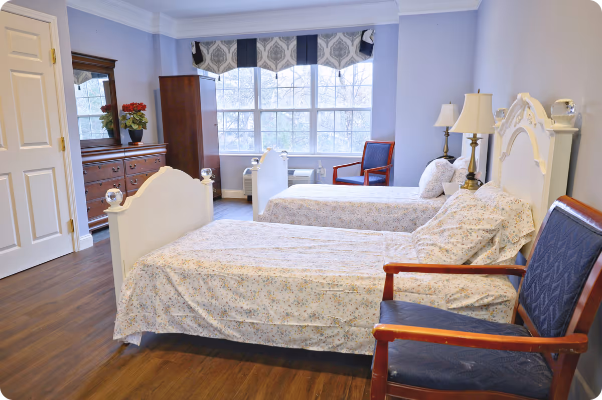 A bright bedroom in an assisted living facility with two twin beds covered in floral bedspreads. The room features wooden flooring, a large window with patterned valance curtains, two wooden armchairs with blue cushions, a wooden dresser with a mirror, and two bedside lamps.