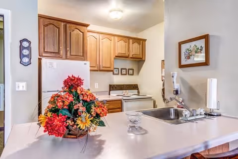 A kitchen area with wooden cabinets, a white refrigerator, and a white stove. A countertop with a sink and faucet is in the foreground, decorated with a colorful flower arrangement and a glass bowl. A framed picture hangs on the wall above the sink.