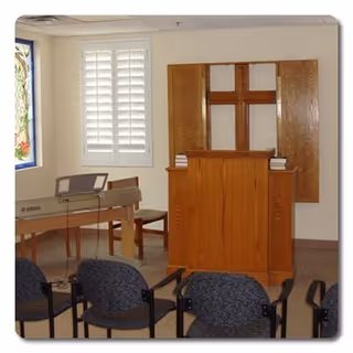Interior of a small chapel or prayer room with a wooden pulpit featuring a cross, a Yamaha keyboard on a stand, several chairs arranged facing the pulpit, and a window with white shutters and stained glass.