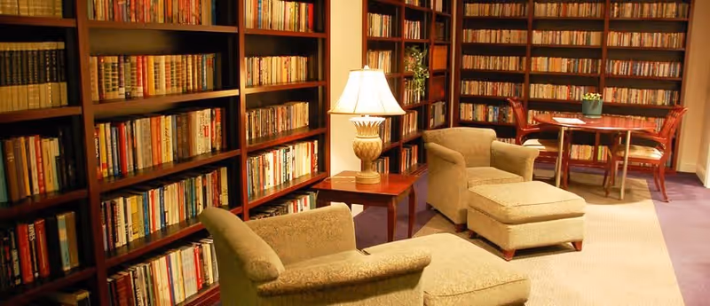 A cozy library room with tall wooden bookshelves filled with books, two upholstered armchairs with matching ottomans, a wooden side table with a decorative lamp, and a round wooden table with chairs in the background.