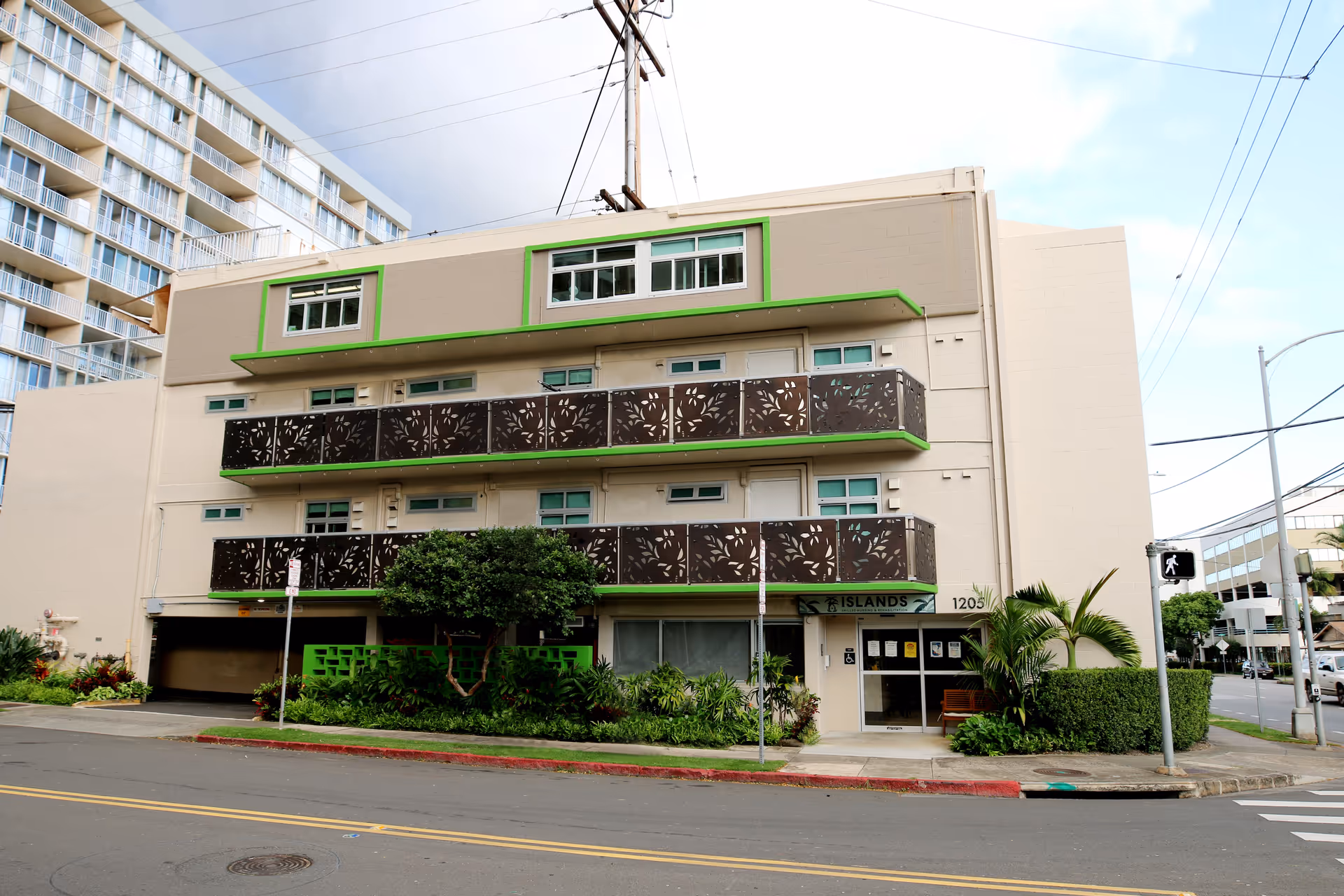 Exterior view of a three-story building with decorative balcony railings and green trim, located on a street corner with some greenery and palm plants near the entrance. The building has a sign that reads 'Islands' near the entrance door.
