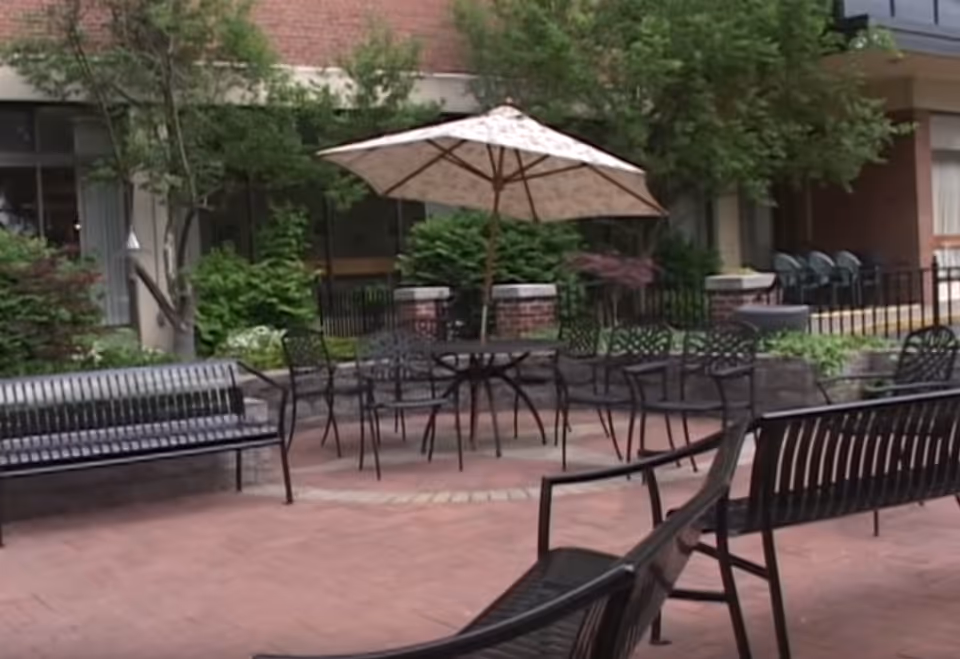 Courtyard patio with metal benches, a table and umbrella, chairs, brick paving and landscaping in front of a building.