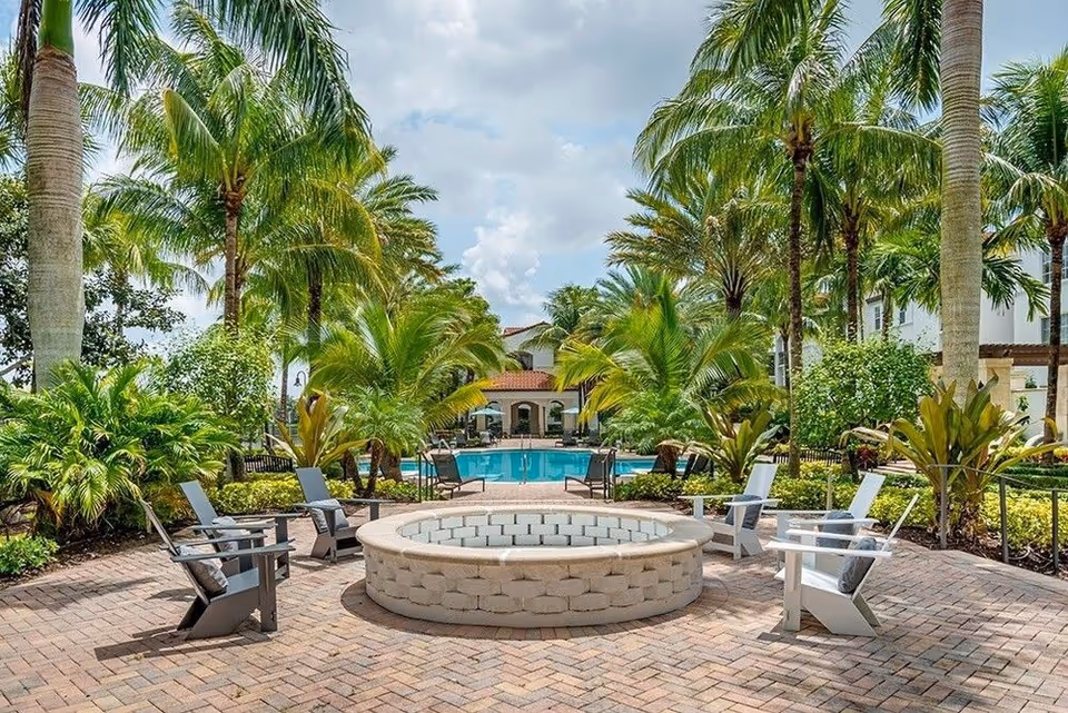 Outdoor seating area with a circular stone fire pit surrounded by six Adirondack chairs on a brick patio, palm trees and lush greenery around, and a swimming pool with lounge chairs in the background under a partly cloudy sky.
