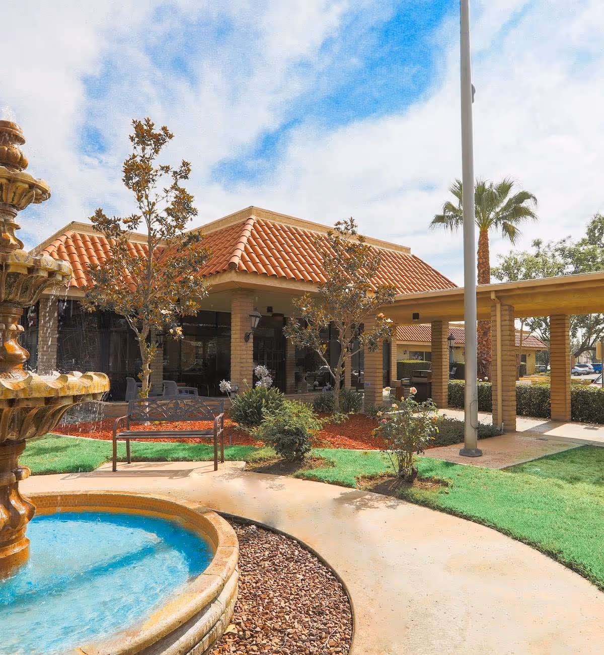 Outdoor view of a senior living facility with a water fountain in the foreground, a curved walkway, green grass, small trees, and a building with a red tiled roof and covered walkway under a partly cloudy sky.