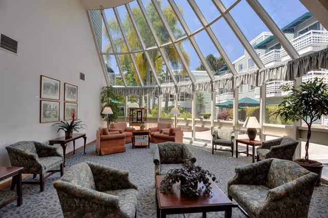 A bright and spacious common area with large floor-to-ceiling windows and a glass ceiling letting in natural light. The room is furnished with multiple upholstered armchairs and sofas arranged around wooden coffee tables. There are several potted plants and table lamps, and outside the windows, greenery and parts of the building exterior are visible.