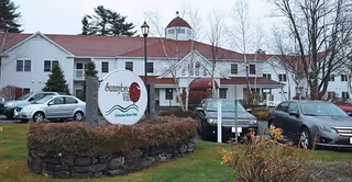 Exterior view of Sunnybrook Senior Living facility showing a large white building with a red roof and a cupola on top. Several cars are parked in front of the building, and a sign with the facility's name is displayed on a stone-bordered grassy area near the parking lot. Trees without leaves are visible in the background.
