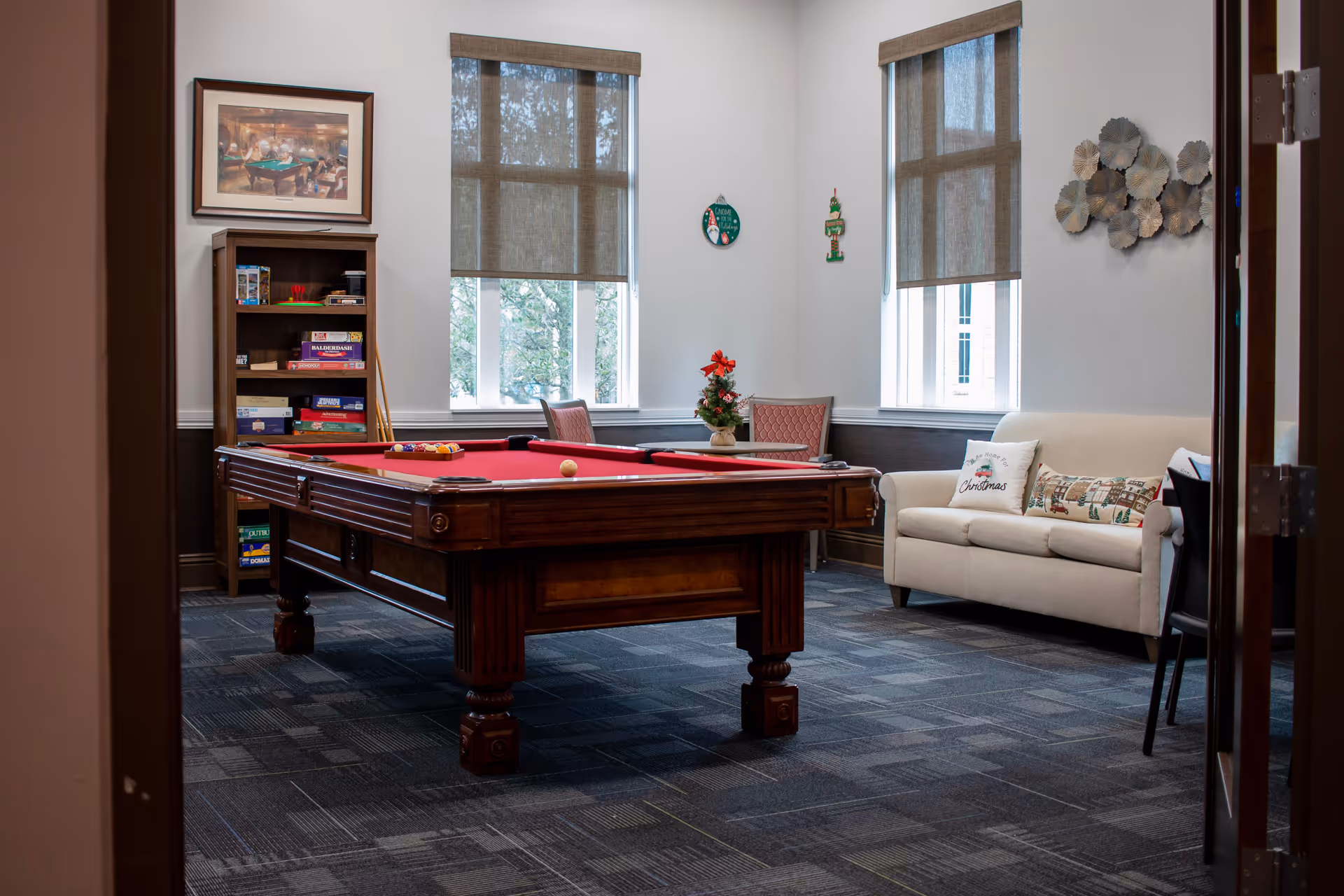 A cozy game room with a red-felt pool table centered, a sofa, bookshelf, and windows.