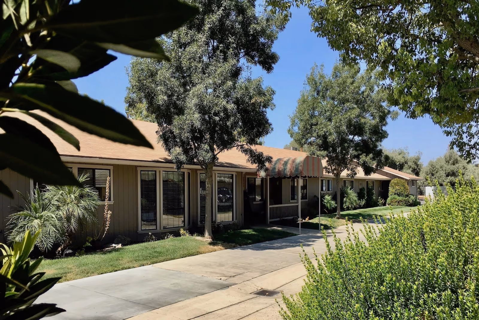 Exterior view of a single-story building with a brown roof and beige walls, surrounded by green trees and bushes under a clear blue sky. A sidewalk runs alongside the building, which has several windows and a small covered entrance with a striped awning.