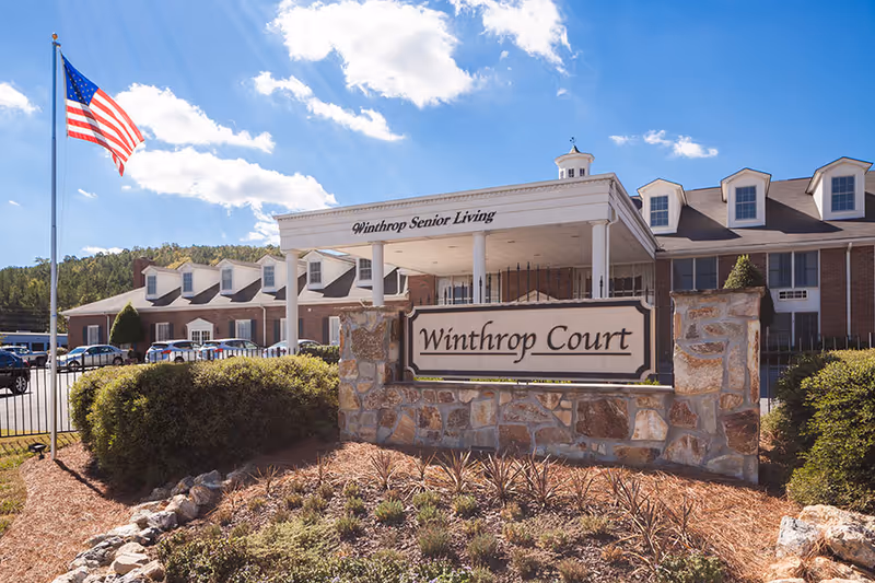 Front exterior of Winthrop Court senior living facility showing the entrance portico, stone sign, American flag, and landscaped grounds.
