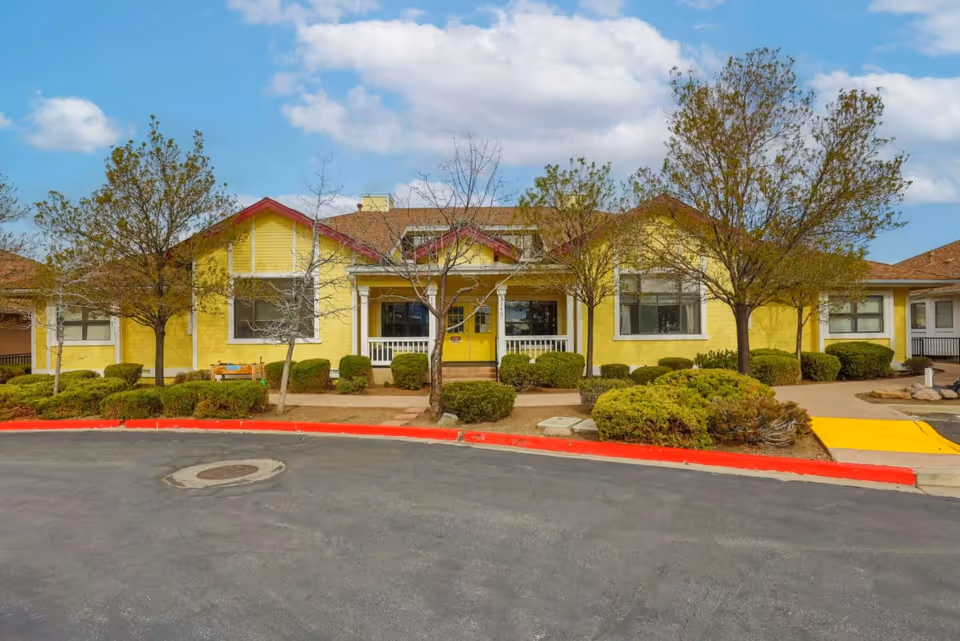 Front exterior view of a single-story yellow building with a red roof and white trim, surrounded by small trees and bushes, under a partly cloudy sky.