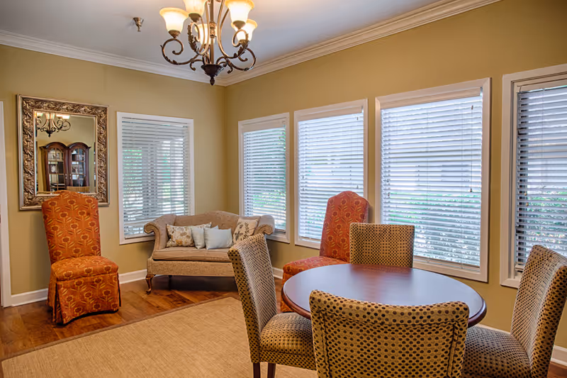 A cozy senior living common area with a round wooden table surrounded by four patterned chairs. There are two orange upholstered chairs and a small beige loveseat with decorative pillows against the wall. The room has multiple windows with white blinds, a large ornate mirror on one wall, and a chandelier hanging from the ceiling. The walls are painted a warm beige color and the floor is wooden with a beige area rug.