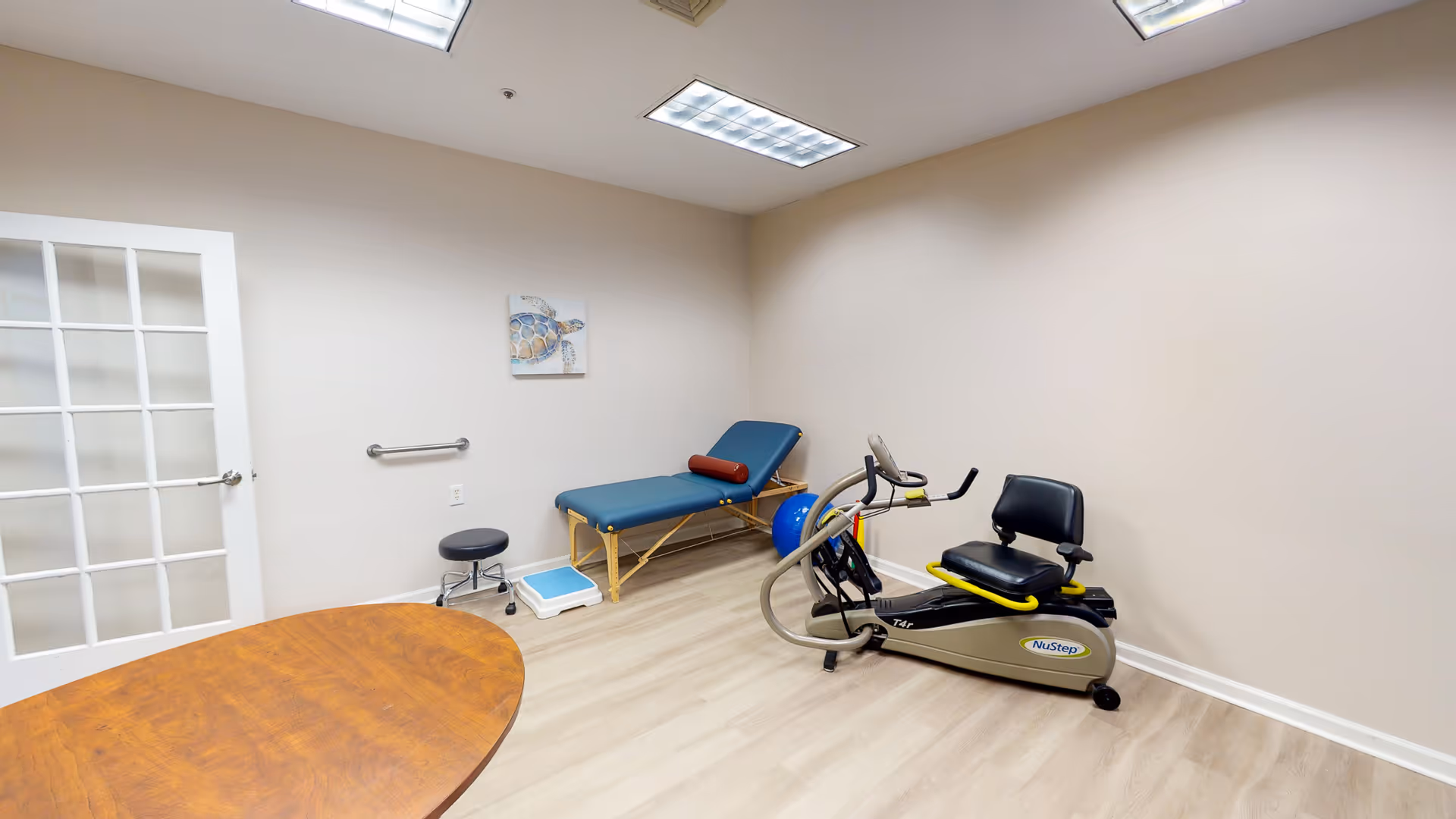 Therapy room with a treatment table, NuStep recumbent exercise bike, stool and round wooden table.