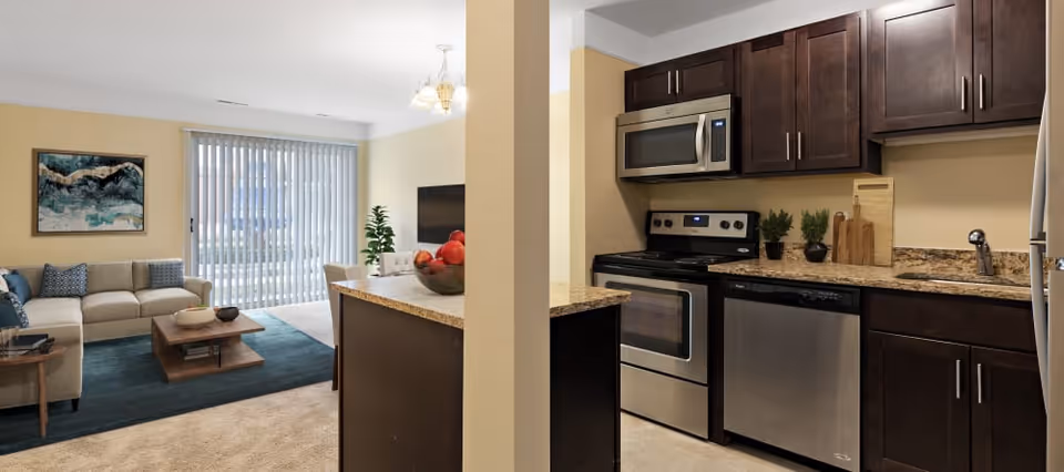 Interior view of a senior living apartment showing a kitchen with stainless steel appliances including a microwave, stove, and dishwasher. The kitchen has dark wood cabinets and granite countertops. Adjacent to the kitchen is a living room area with a beige sofa, a wooden coffee table, a blue rug, a side table, a wall-mounted TV, and a large window with vertical blinds letting in natural light.