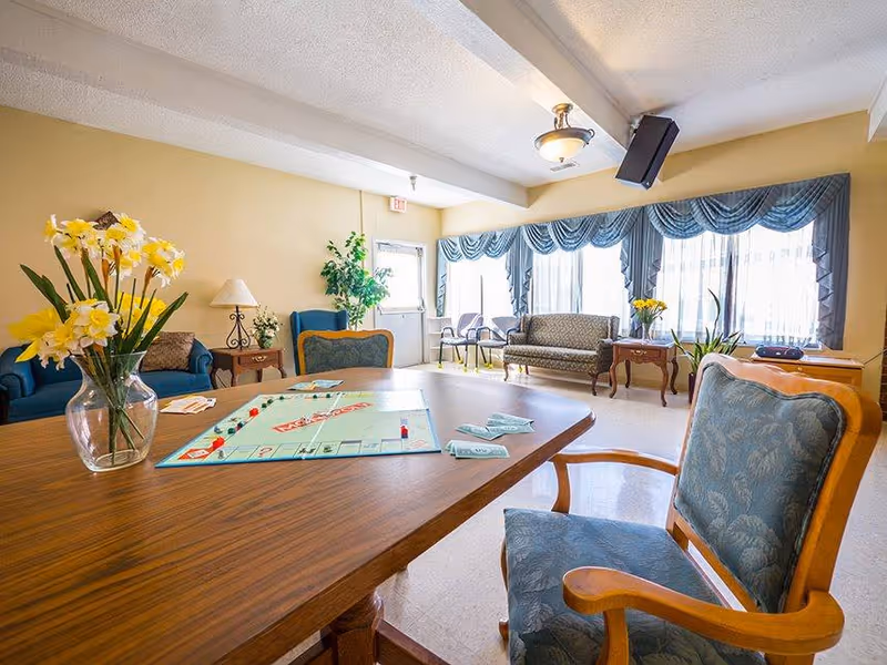 A bright and cozy common area in a senior living facility with a wooden table in the foreground featuring a Monopoly board game set up. The room has comfortable upholstered chairs and sofas, a vase with yellow flowers on the table, large windows with blue curtains allowing natural light, and a potted plant near the door.