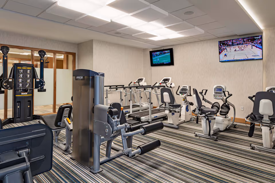 A fitness room with various exercise equipment including treadmills, recumbent bikes, and strength training machines. Two wall-mounted televisions are showing sports games. The room has striped carpeting and neutral-colored walls with a drop ceiling and fluorescent lighting.