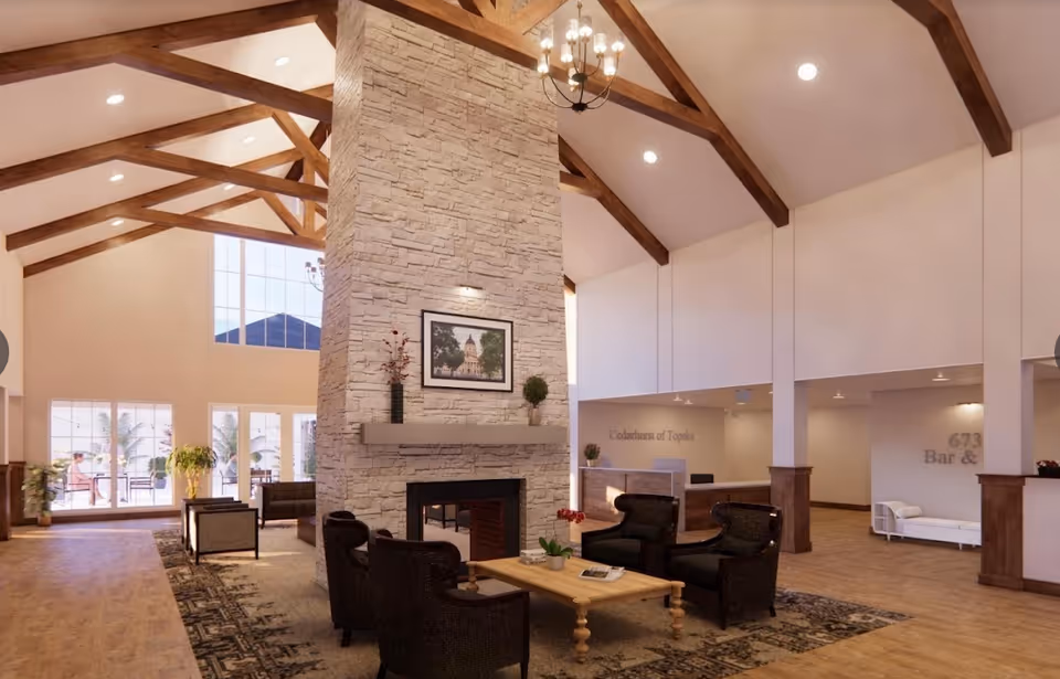 Spacious senior living facility common area with high vaulted ceilings and exposed wooden beams. A large stone fireplace stands centrally with a framed picture above the mantle. Comfortable black armchairs and a wooden coffee table are arranged around the fireplace on a patterned rug. Large windows and glass doors allow natural light to fill the room. The reception desk is visible in the background with the text 'Cedarhurst of Topeka' on the wall behind it. There are plants and additional seating areas near the windows.