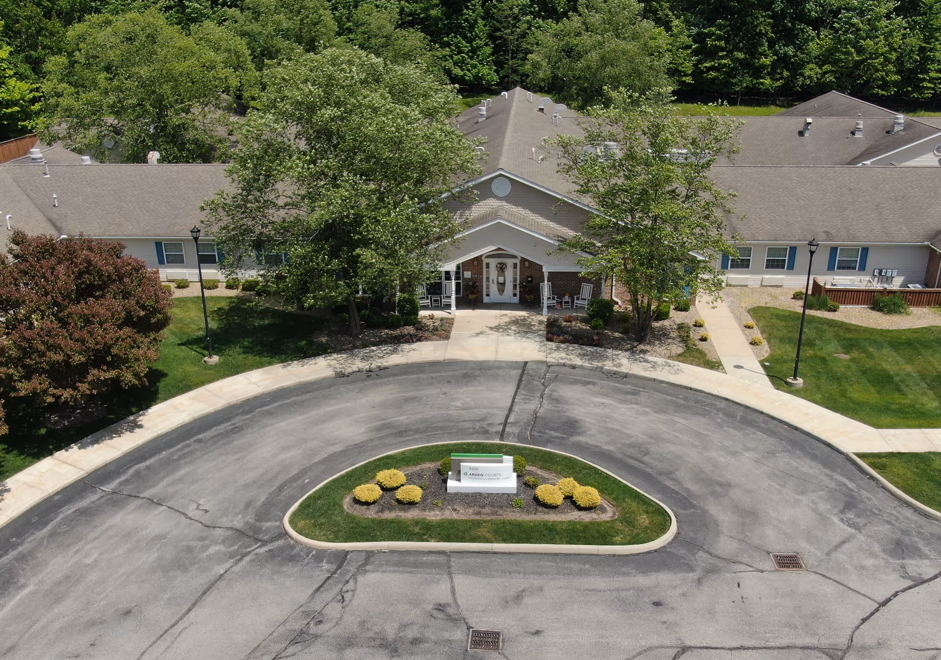 Aerial view of the front entrance of Arden Courts - ProMedica Memory Care Community in Parma, showing a circular driveway with a landscaped island in the center, trees, and a single-story building with a covered porch and rocking chairs.