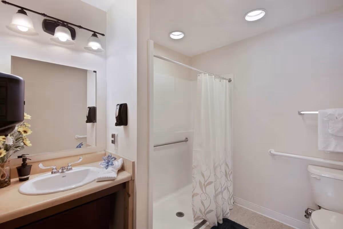A clean and well-lit bathroom with a sink and countertop on the left, a large mirror above the sink, a soap dispenser mounted on the wall, and a vase with flowers. To the right is a shower area with a white curtain featuring a subtle leaf pattern, grab bars for safety, and a toilet with a towel hanging on a rack above it. The walls are painted white, and the ceiling has recessed lighting.